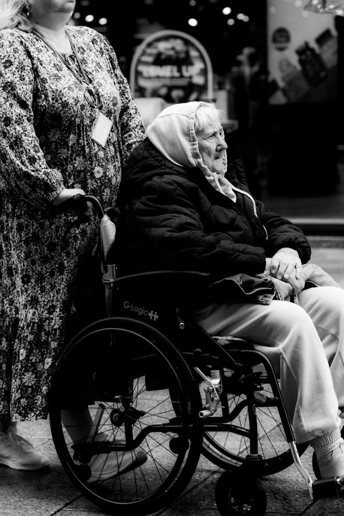 Black and white photo of an elderly woman in a wheelchair pushed by a caregiver on a street in Dublin. Urban life scene.